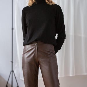 May include: A person wearing a black turtleneck sweater and brown leather pants. The pants have a classic, straight-leg fit. The outfit is simple and stylish, suitable for various occasions.