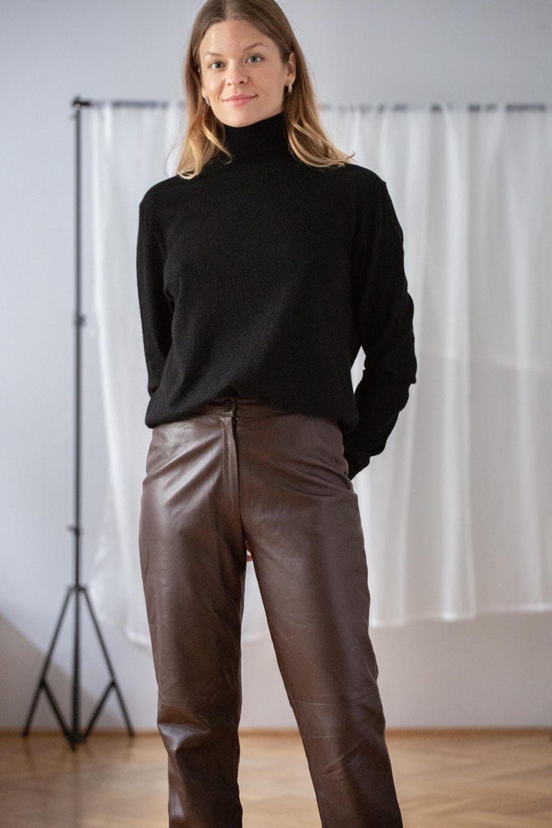 May include: A person wearing a black turtleneck sweater and brown leather pants. The pants have a classic, straight-leg fit. The outfit is simple and stylish, suitable for various occasions.