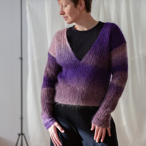 Può includere: Un maglione con scollo a V nei toni del viola e malva. Il maglione ha un effetto sfumato, che passa dal malva chiaro in alto al viola scuro in basso. Le maniche sono lunghe e il materiale appare morbido e soffice. La persona indossa pantaloni neri.