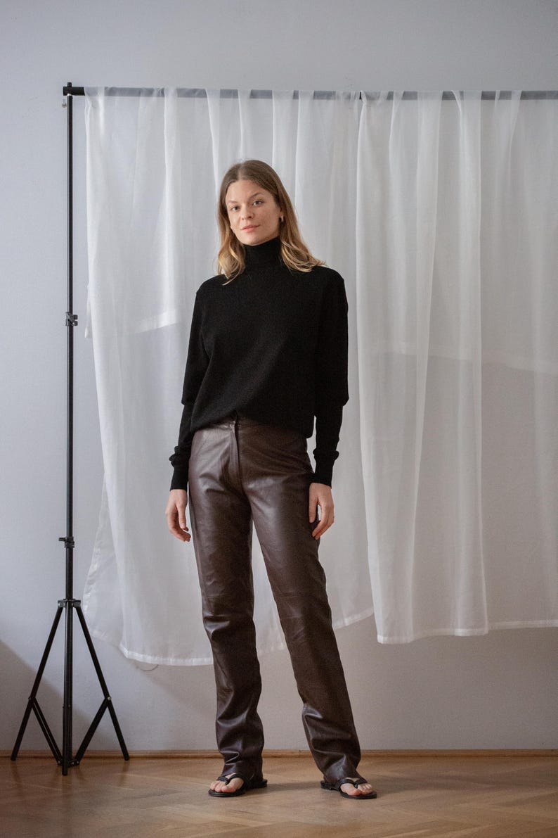 May include: A person wearing a black turtleneck sweater and brown leather pants. The outfit is paired with black sandals. The background features a white curtain and a black tripod.