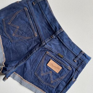 May include: Dark blue denim shorts with a rolled hem. The shorts feature two back pockets with the Wrangler logo stitched in a contrasting tan thread. The Wrangler logo is also on a leather patch on the back pocket.