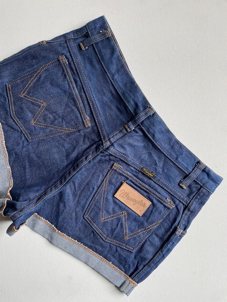 May include: Dark blue denim shorts with a rolled hem. The shorts feature two back pockets with the Wrangler logo stitched in a contrasting tan thread. The Wrangler logo is also on a leather patch on the back pocket.