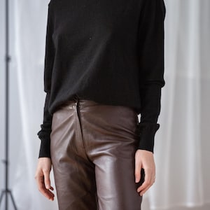 May include: A black turtleneck sweater paired with brown leather pants. The sweater has long sleeves and a ribbed cuff. The pants are a rich brown color with a classic fit. The outfit is simple and elegant.