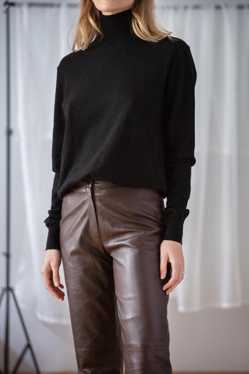 May include: A black turtleneck sweater paired with brown leather pants. The sweater has long sleeves and a ribbed cuff. The pants are a rich brown color with a classic fit. The outfit is simple and elegant.