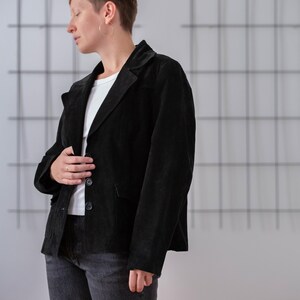 May include: A person wearing a black suede jacket with a white t-shirt underneath. They are wearing dark wash jeans and have short, dark hair.