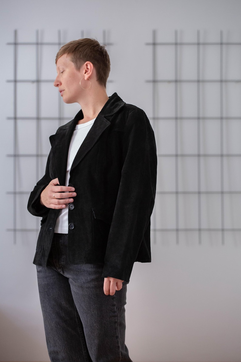 May include: A person wearing a black suede jacket with a white t-shirt underneath. They are wearing dark wash jeans and have short, dark hair.
