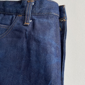 May include: Close-up of dark blue denim jeans. The jeans have a classic five-pocket design with a button and zipper closure. The stitching is a contrasting tan color, and the fabric has a slightly worn appearance.