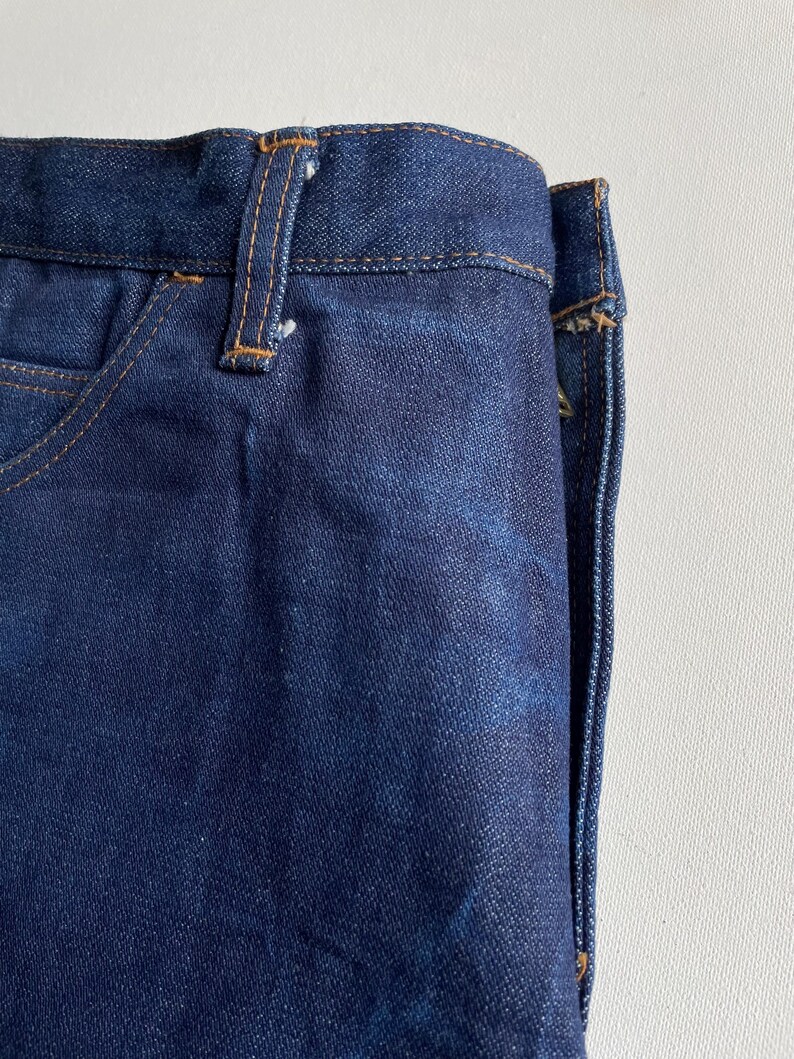 May include: Close-up of dark blue denim jeans. The jeans have a classic five-pocket design with a button and zipper closure. The stitching is a contrasting tan color, and the fabric has a slightly worn appearance.
