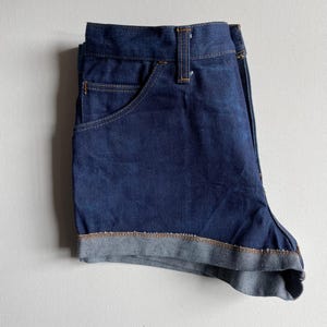 May include: Dark blue denim shorts with a classic five-pocket design. The shorts have a folded hem and feature contrasting tan stitching. The shorts are folded neatly, showcasing the front pocket and the folded hem detail.