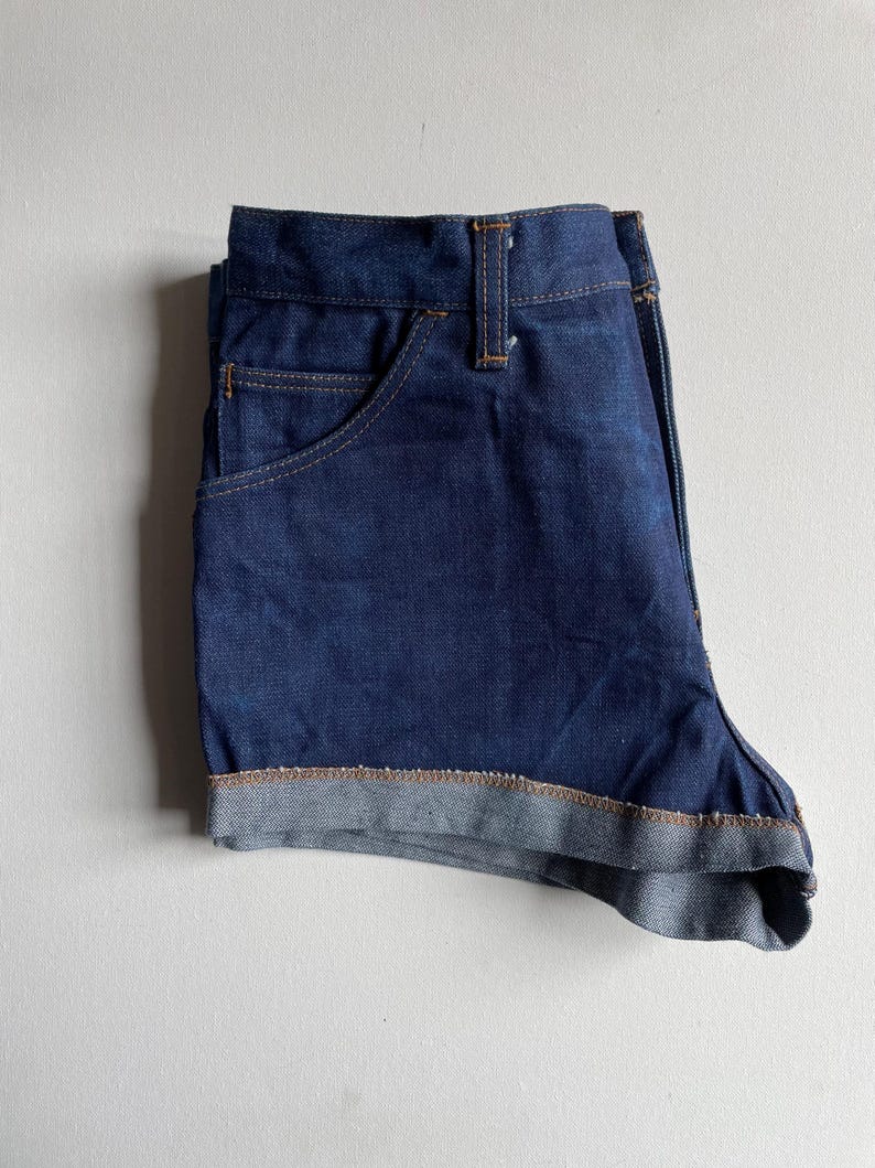 May include: Dark blue denim shorts with a classic five-pocket design. The shorts have a folded hem and feature contrasting tan stitching. The shorts are folded neatly, showcasing the front pocket and the folded hem detail.
