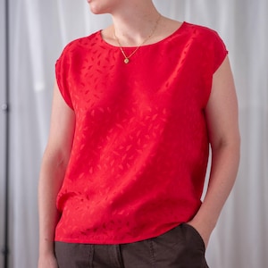 May include: A bright red, short-sleeved top with a subtle leaf pattern. The top has a round neckline and is paired with brown pants. A gold necklace with a small pendant is visible.