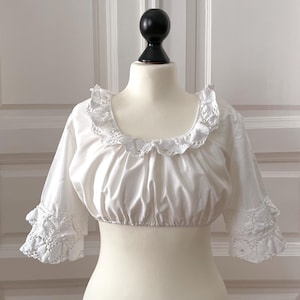 May include: A white, cropped blouse with a square neckline and short sleeves. The neckline and sleeve cuffs feature delicate lace trim. The blouse is displayed on a mannequin.