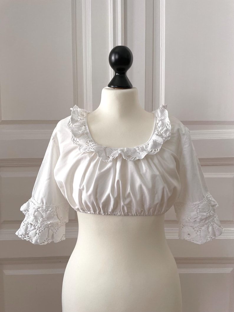 May include: A white, cropped blouse with a square neckline and short sleeves. The neckline and sleeve cuffs feature delicate lace trim. The blouse is displayed on a mannequin.