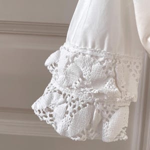 May include: Close-up of a white sleeve with a crochet cuff. The cuff features a detailed, openwork design with a scalloped edge. The fabric appears to be a soft, lightweight material.