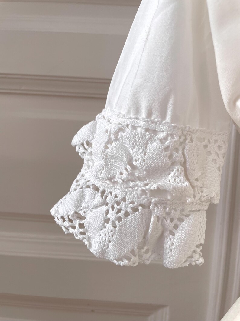 May include: Close-up of a white sleeve with a crochet cuff. The cuff features a detailed, openwork design with a scalloped edge. The fabric appears to be a soft, lightweight material.