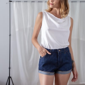 May include: A woman wearing a white sleeveless top with a cowl neck and dark blue denim shorts. The shorts have rolled-up cuffs and pockets. The outfit is casual and stylish.