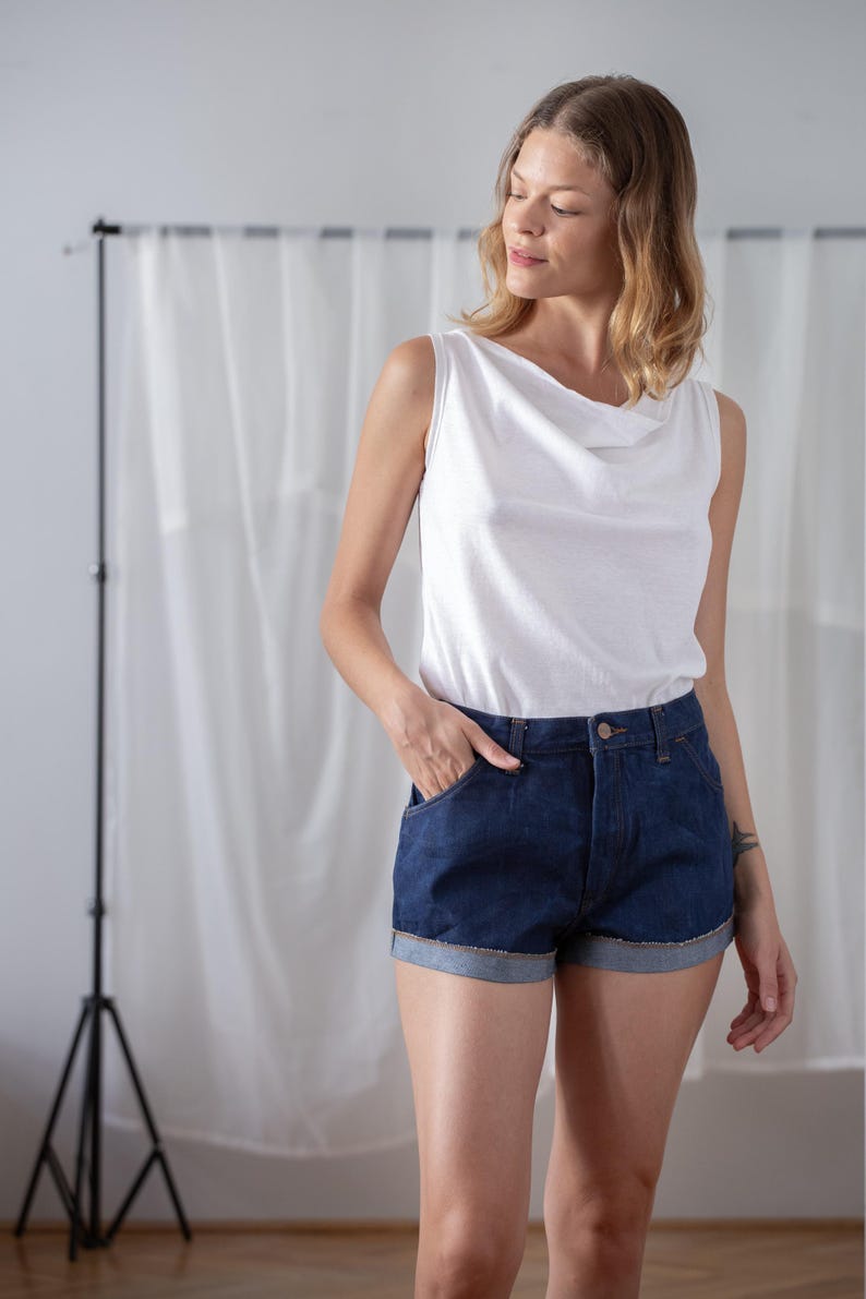 May include: A woman wearing a white sleeveless top with a cowl neck and dark blue denim shorts. The shorts have rolled-up cuffs and pockets. The outfit is casual and stylish.