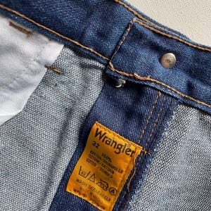 May include: Close-up of blue denim jeans with a Wrangler label. The label is yellow with black text indicating size 32 and 100% cotton. The jeans have brown stitching and a silver button.