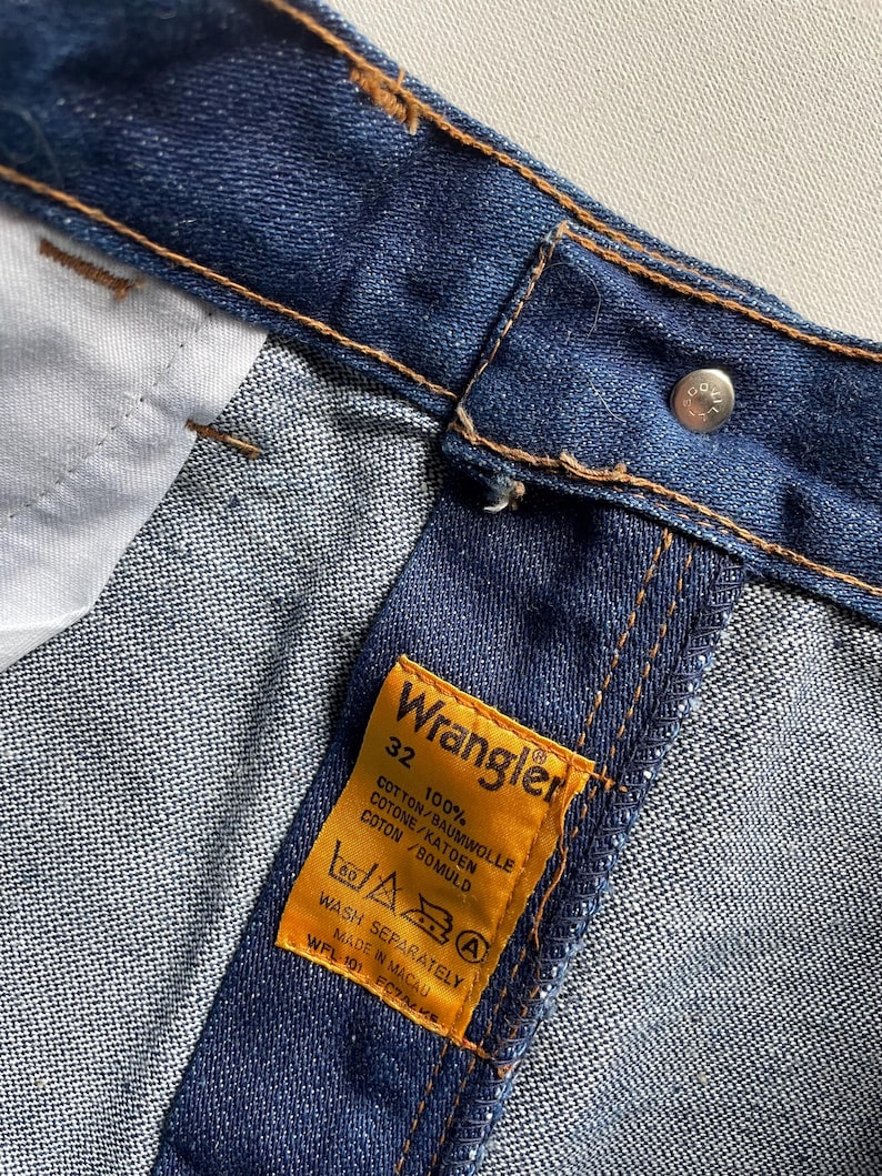 May include: Close-up of blue denim jeans with a Wrangler label. The label is yellow with black text indicating size 32 and 100% cotton. The jeans have brown stitching and a silver button.
