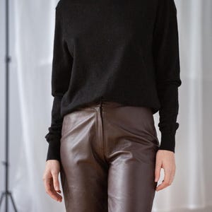 May include: A woman wearing a black turtleneck sweater and brown leather pants. The sweater has long sleeves and a high neckline. The pants are a dark brown color and have a fitted silhouette. The outfit is simple and classic.