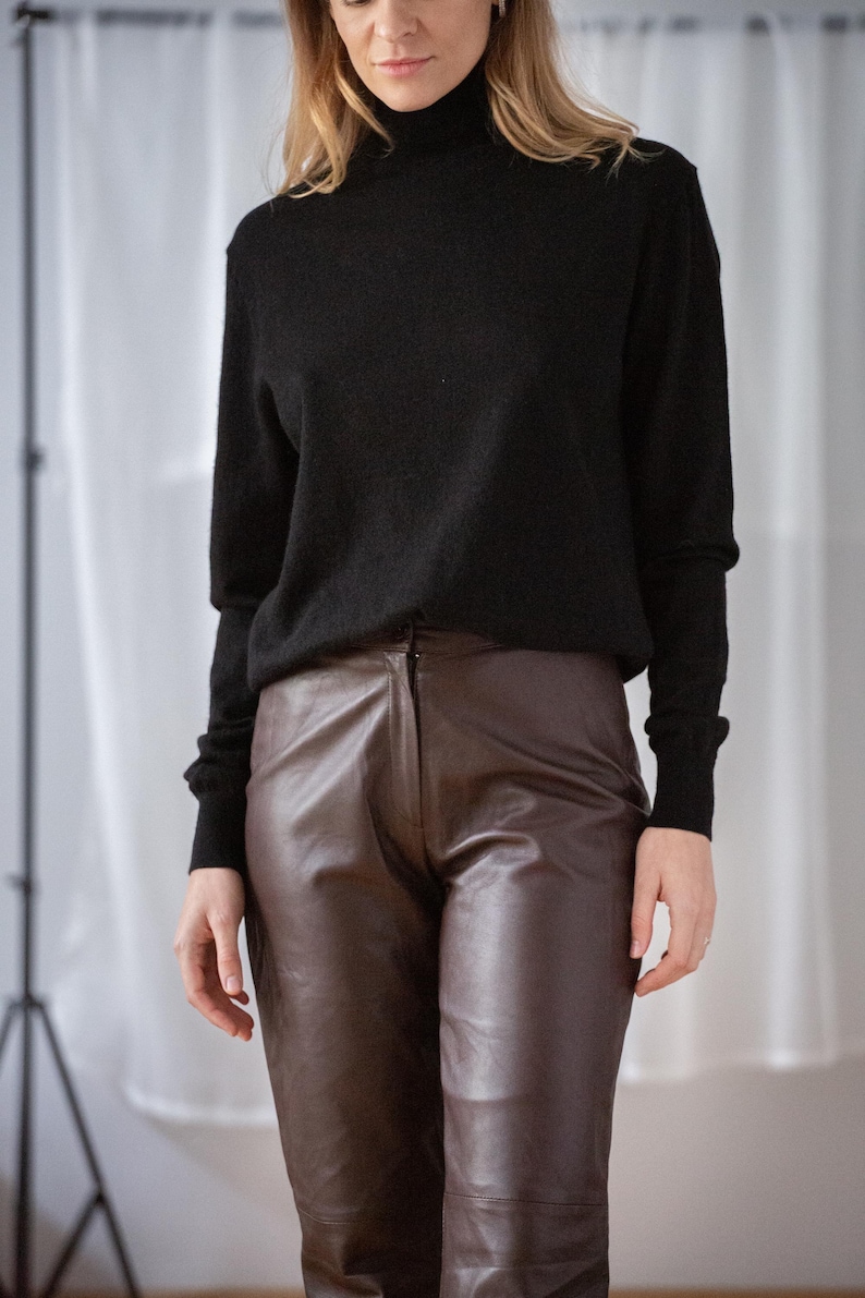 May include: A woman wearing a black turtleneck sweater and brown leather pants. The sweater has long sleeves and a high neckline. The pants are a dark brown color and have a fitted silhouette. The outfit is simple and classic.