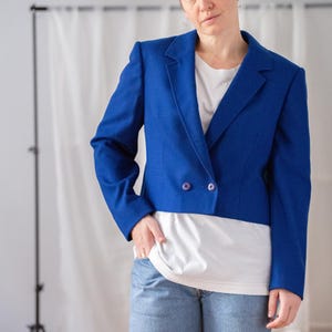 Op de afbeelding: Een korte, koningsblauwe blazer met een dubbele rij knopen en twee paarse knopen. De blazer wordt gedragen over een wit t-shirt en een lichtblauwe jeans. Een eenvoudige en stijlvolle outfit.