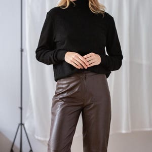 May include: A woman wearing a black turtleneck sweater and brown leather pants. The outfit is simple and classic, with a focus on texture and neutral colors. The pants have a high waist and a straight leg.