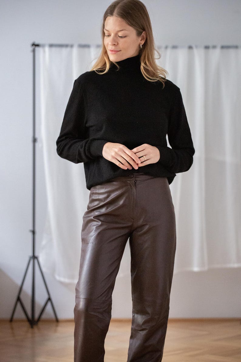 May include: A woman wearing a black turtleneck sweater and brown leather pants. The outfit is simple and classic, with a focus on texture and neutral colors. The pants have a high waist and a straight leg.