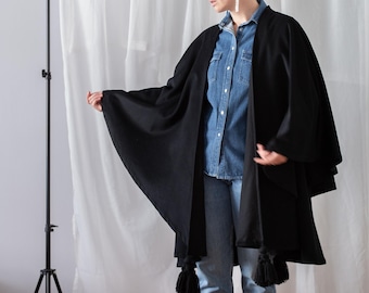 May include: A black open-front cape featuring a smooth fabric construction with large black tassels attached to the bottom corners. The garment is worn over a blue denim button-down shirt, light blue denim jeans, and black leather ankle boots.