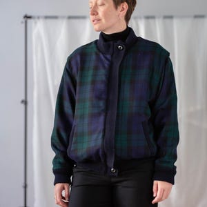 Könnte beinhalten: Eine karierte Bomberjacke in Grün und Marineblau. Die Jacke hat einen gerippten Kragen, Bündchen und Saum. Sie hat einen Knopfverschluss und zwei Vordertaschen. Die Jacke wird mit einer schwarzen Hose und einem schwarzen Rollkragenpullover getragen.