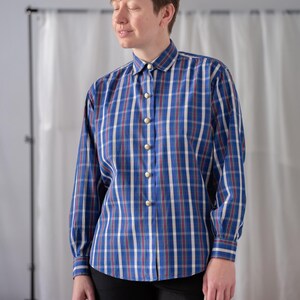 May include: A long-sleeved plaid shirt in blue, white, and red. The shirt has a button-down front with gold-colored buttons and a classic collar. The plaid pattern is a grid of vertical and horizontal lines.