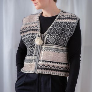 Peut inclure: Une femme portant un chemisier noir à manches longues et un pantalon noir avec un gilet en tricot à motifs gris, blanc et noir, avec une fermeture éclair et un pompon.