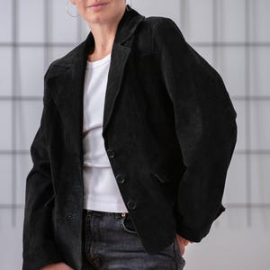 May include: A person wearing a black suede blazer with a white t-shirt and dark wash jeans.