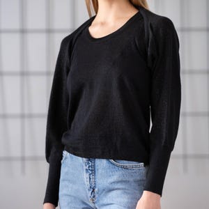 May include: A black sweater with a round neckline and long sleeves. The sweater has a subtle shimmer and is paired with light blue jeans. The sleeves have a slight puff at the shoulders.