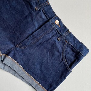 May include: Dark blue denim shorts with a classic design. The shorts feature a button closure, front pockets, and rolled-up hems. The stitching is a contrasting tan color, and the overall look is casual and stylish.