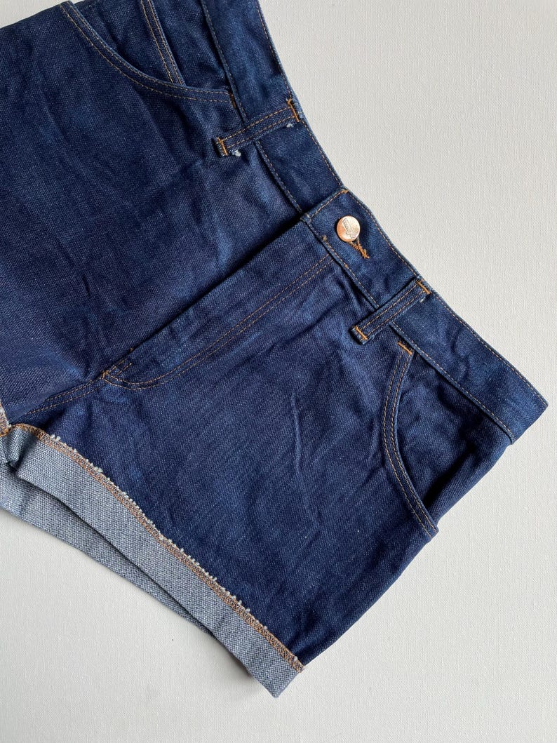 May include: Dark blue denim shorts with a classic design. The shorts feature a button closure, front pockets, and rolled-up hems. The stitching is a contrasting tan color, and the overall look is casual and stylish.