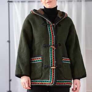 May include: A dark green hooded coat with wooden toggles and colourful trim. The coat features a black trim and a faux fur-lined hood. The colourful trim includes red, green, and white zig-zag patterns. The coat is worn over a black turtleneck.