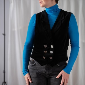 Può includere: Una persona indossa un gilet di velluto nero con bottoni argentati sopra una maglietta a collo alto blu e jeans neri.