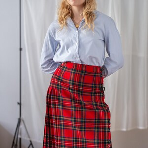 May include: A red tartan kilt with green and yellow checks is paired with a light blue shirt. The kilt, a traditional garment, is fastened with straps. The shirt has a classic collar and buttoned cuffs. The kilt's length is approximately 76 cm.