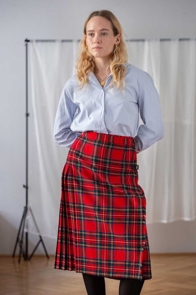 May include: A red tartan kilt with green and yellow checks is paired with a light blue shirt. The kilt, a traditional garment, is fastened with straps. The shirt has a classic collar and buttoned cuffs. The kilt's length is approximately 76 cm.