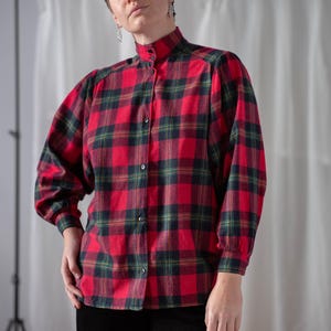 May include: A red, green, and black plaid flannel shirt with a high collar and long sleeves. The shirt is buttoned up and worn with black corduroy pants. The shirt has a classic, vintage style.