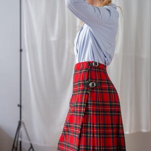 May include: A red tartan skirt with black leather buckle fastenings. The skirt displays a traditional plaid pattern in red, green, and yellow. The skirt is worn with a light blue button-down shirt.