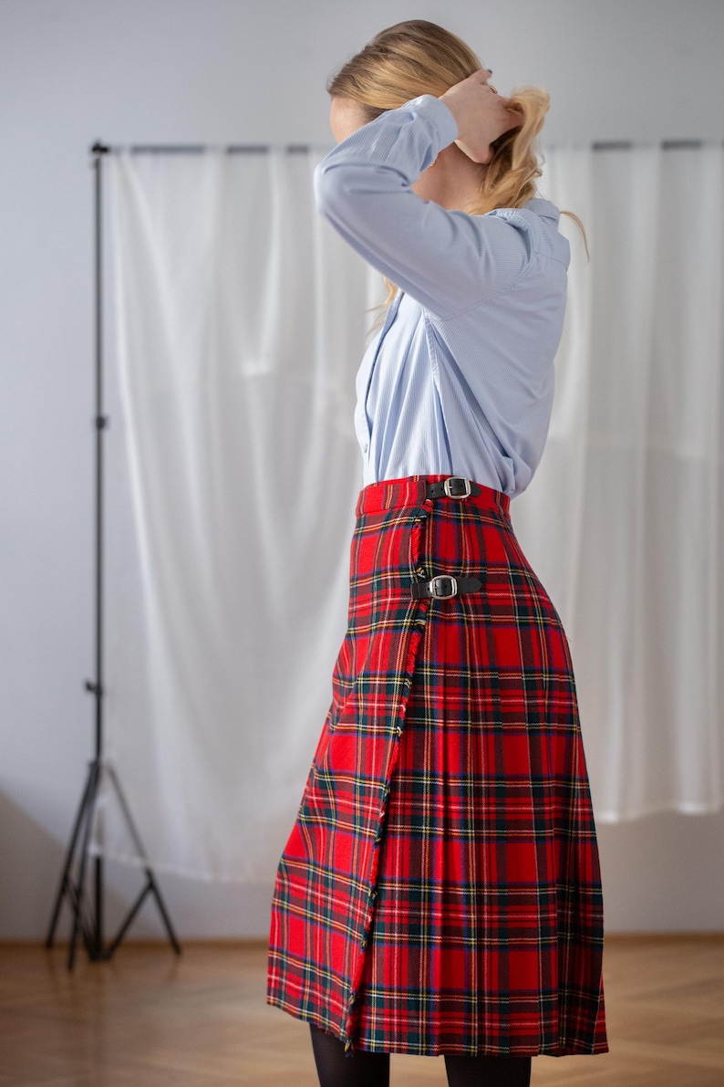 May include: A red tartan skirt with black leather buckle fastenings. The skirt displays a traditional plaid pattern in red, green, and yellow. The skirt is worn with a light blue button-down shirt.