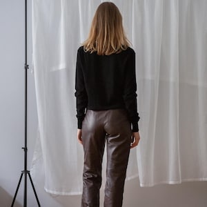 May include: A person wearing a black long-sleeve shirt and brown leather pants. The person is standing in front of a white curtain. The outfit is simple and classic.