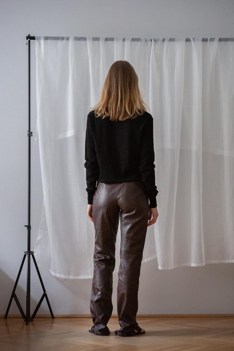 May include: A person wearing a black long-sleeve shirt and brown leather pants. The person is standing in front of a white curtain. The outfit is simple and classic.