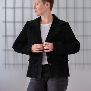 May include: A black suede jacket with a button fastening and two pockets. The jacket is being worn by a person with short brown hair.