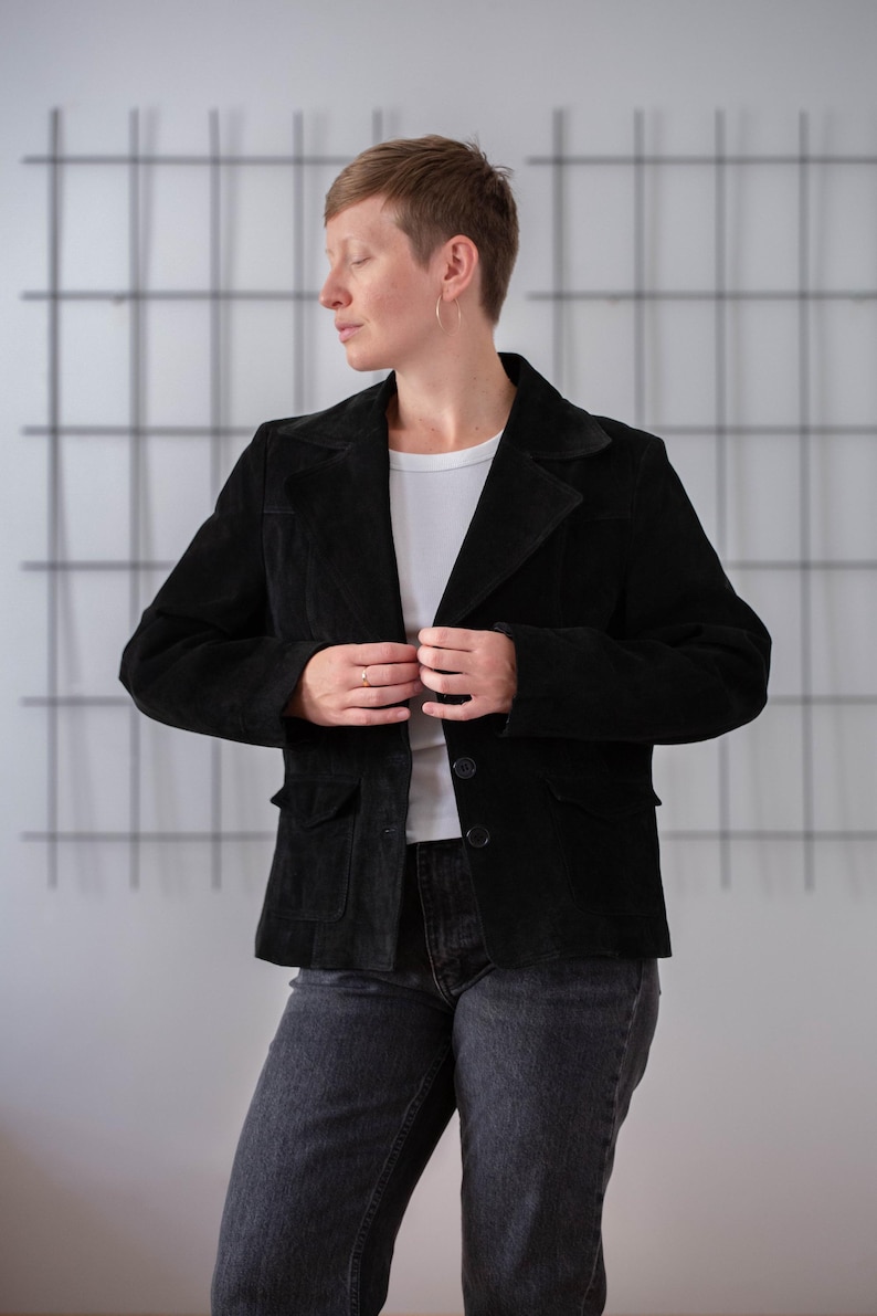 May include: A black suede jacket with a button fastening and two pockets. The jacket is being worn by a person with short brown hair.