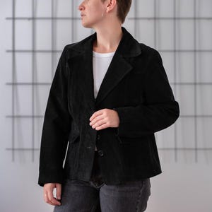 May include: A black suede jacket with a button closure and two pockets. The jacket has a collar and is worn over a white t-shirt.