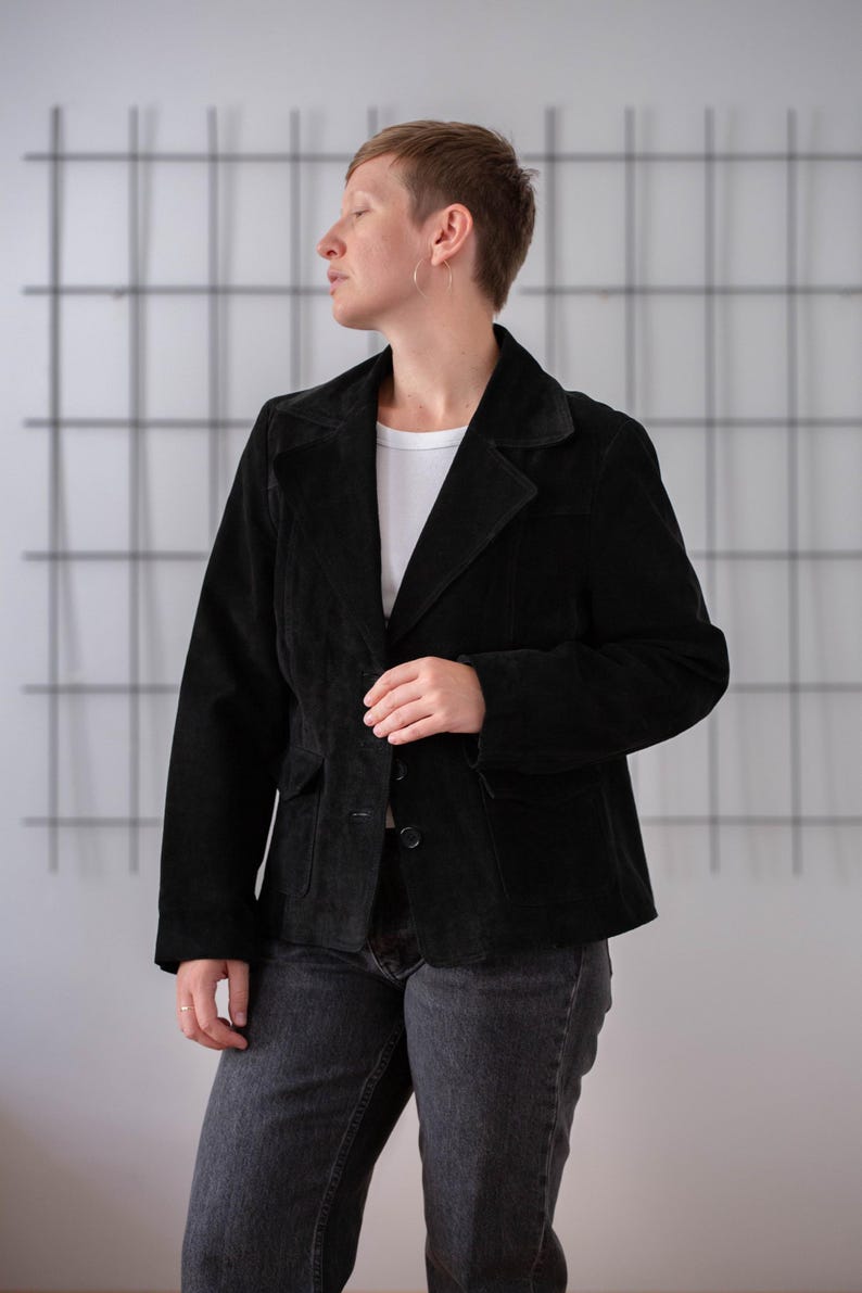 May include: A black suede jacket with a button closure and two pockets. The jacket has a collar and is worn over a white t-shirt.