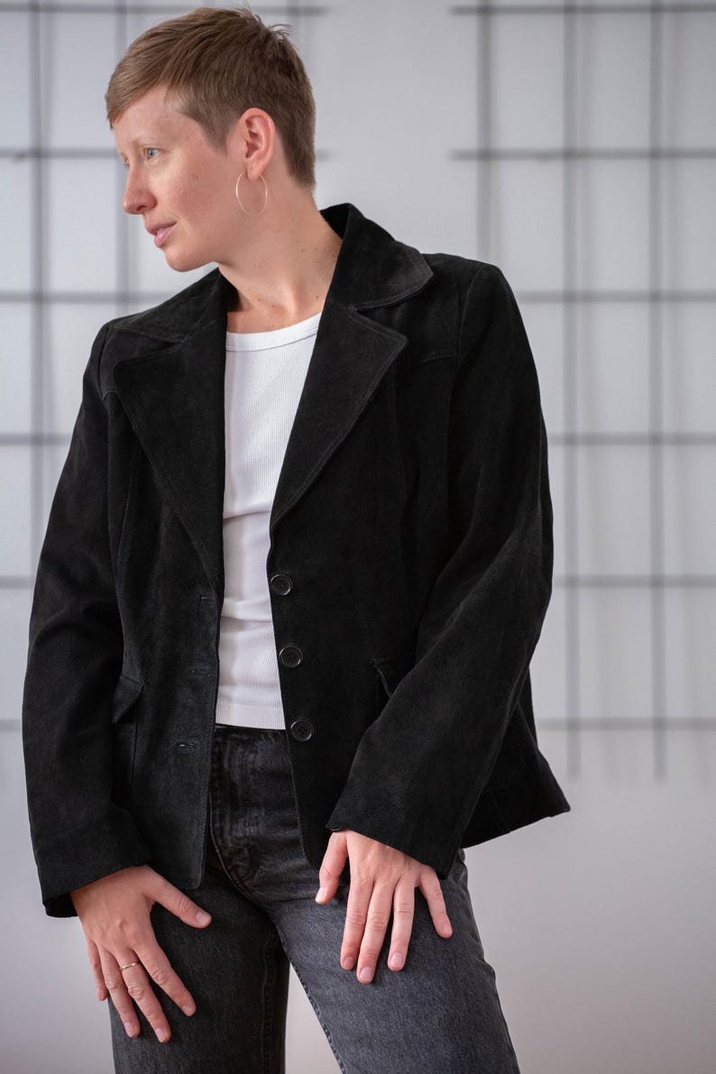 May include: A black suede jacket with a button fastening. The jacket has a collar and two pockets. The person wearing the jacket is standing in front of a white wall.